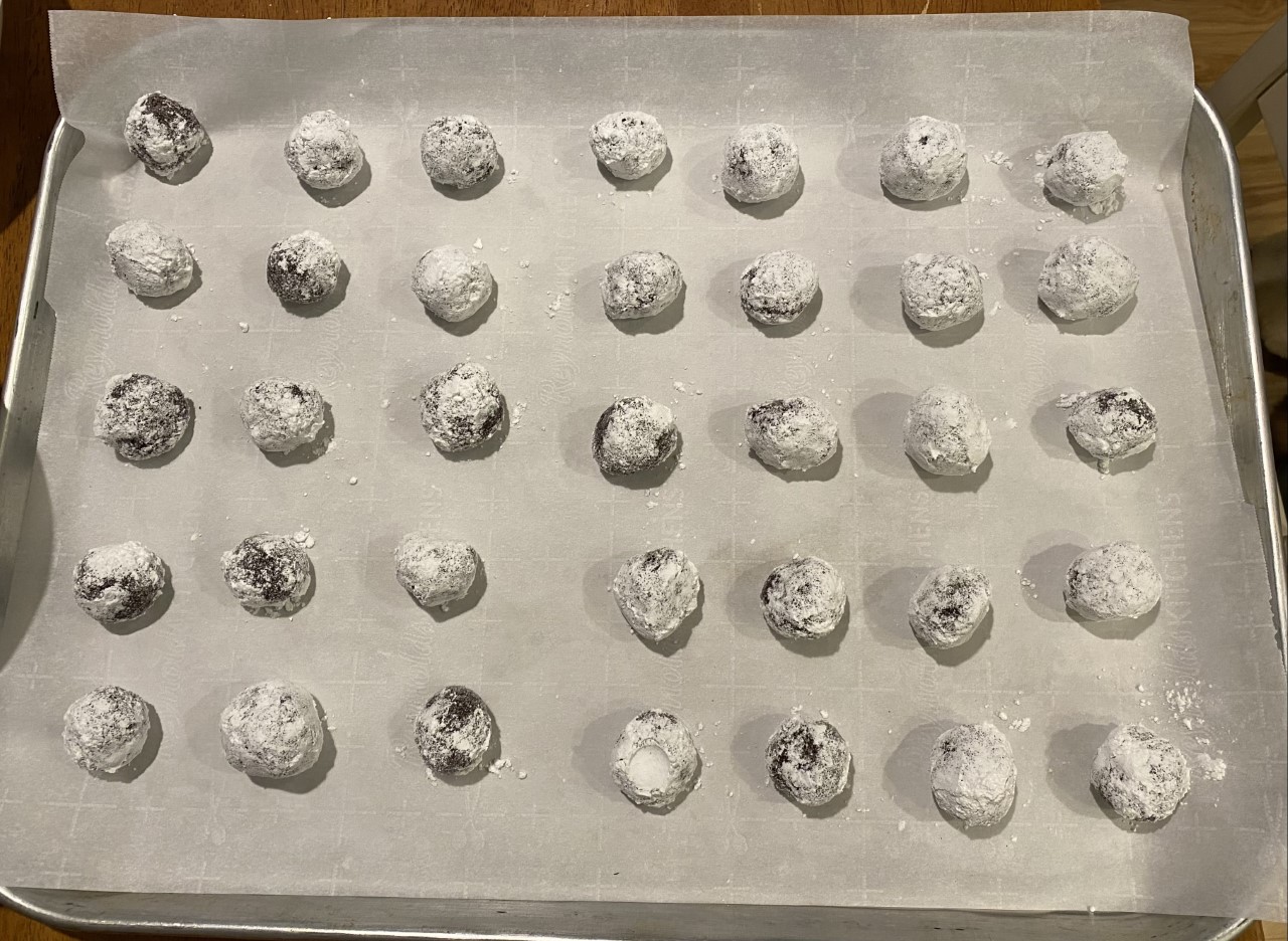 Balls of fudgey cookie dough coated in granulated sugar and confectioners sugar. The balls of dough have been placed on a parchment paper lined cookie sheet approximately two inches apart.