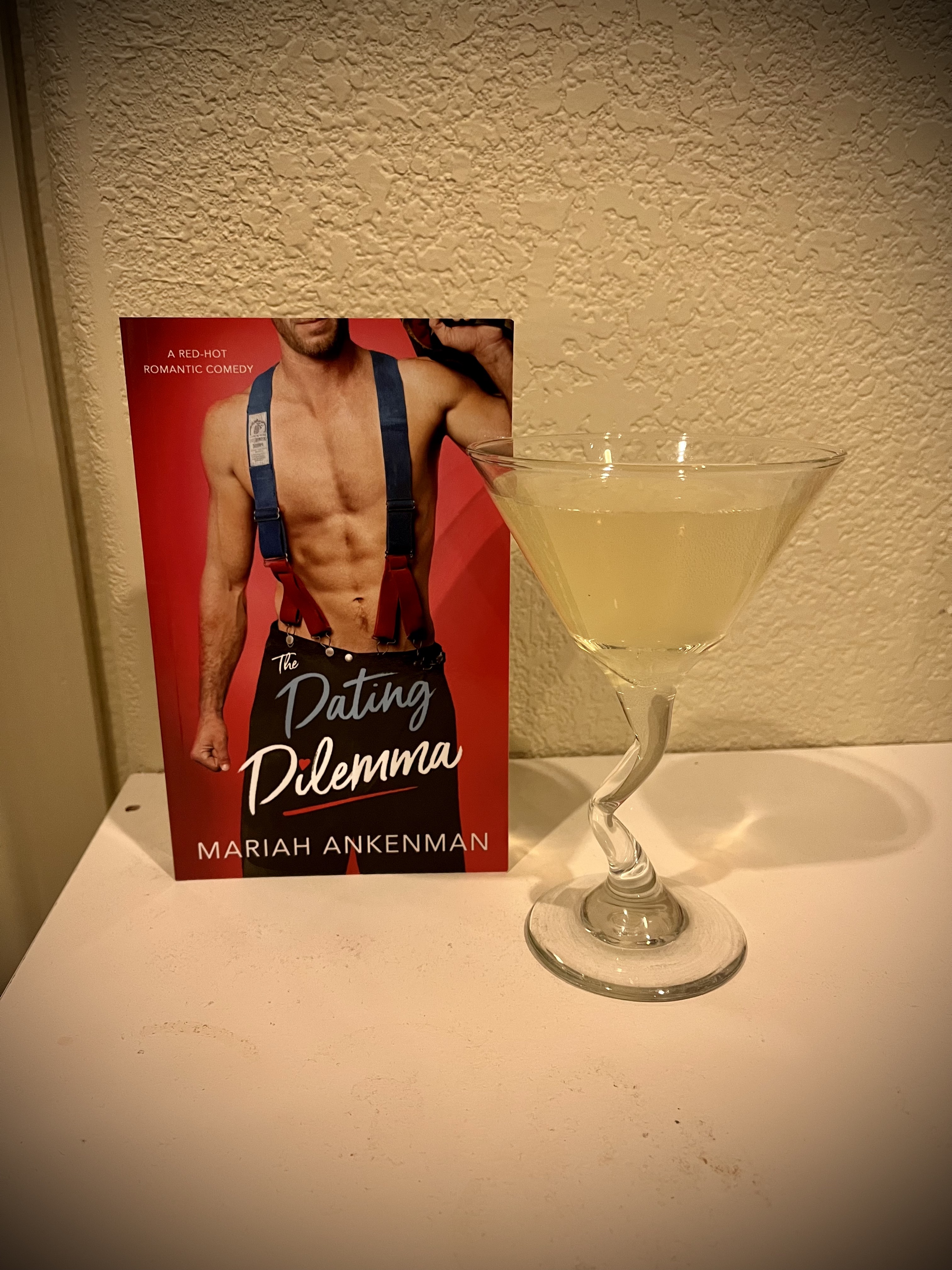 The cover of The Dating Dilemma by Mariah Ankenman sits next to the station 42 cocktail, poured into a martini glass.