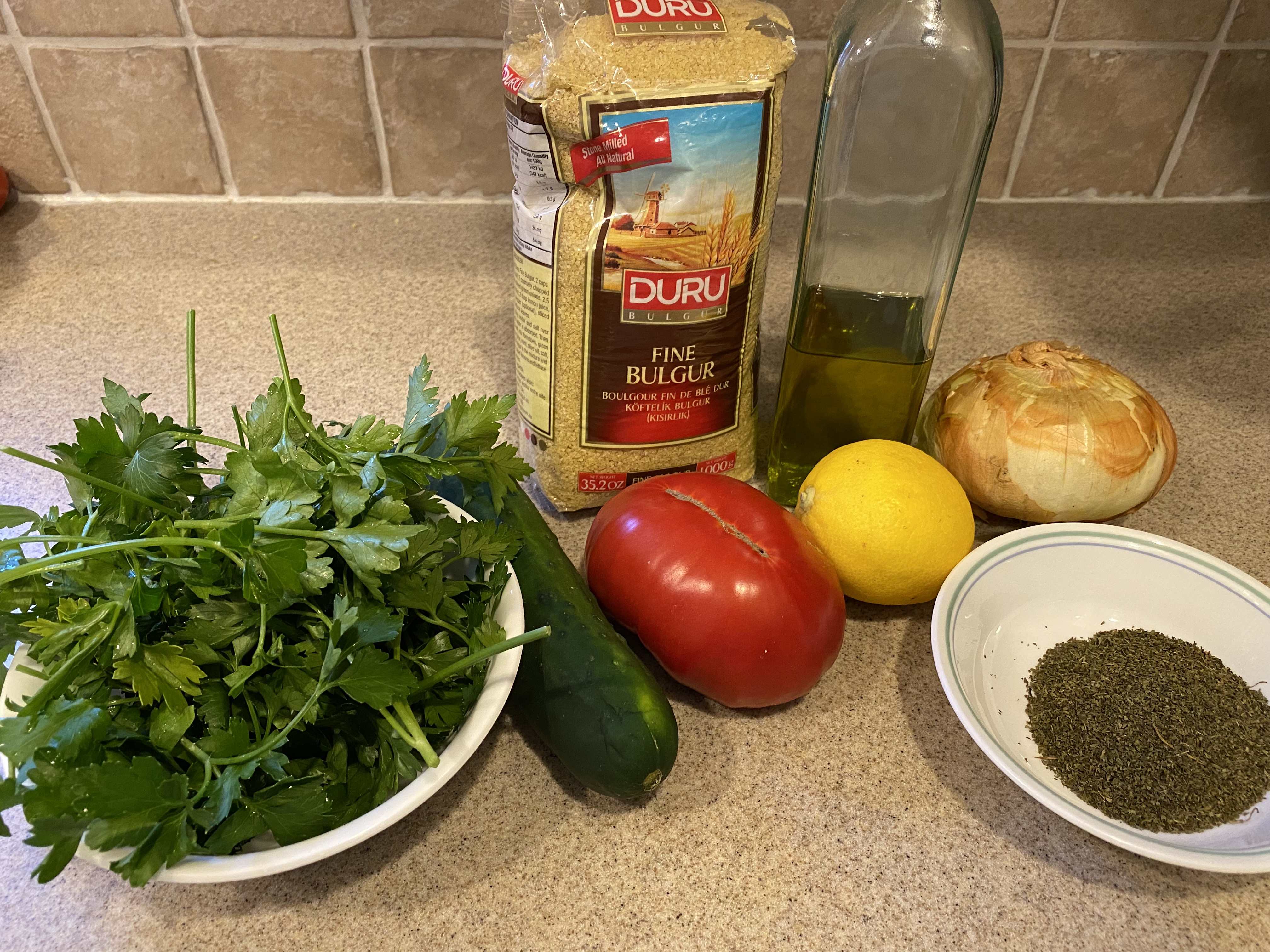 A bag of Duru bran fine bulgur, a bottle of extra virgin olive oil, an onion, a bowl of fresh parsley, a cucumber, a tomato, a lemon, and a bowl of dried, crushed mint, salt, and pepper on a kitchen counter.