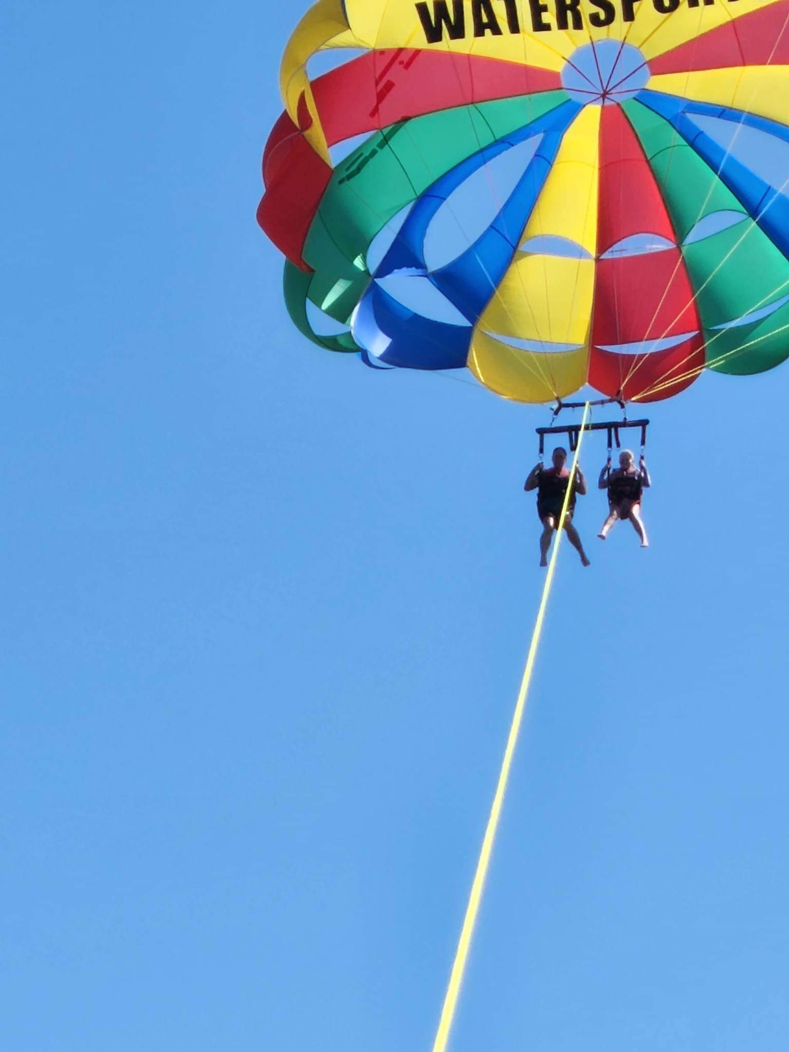 two people are safely harnessed to a parasail against a perfectly blue sky.