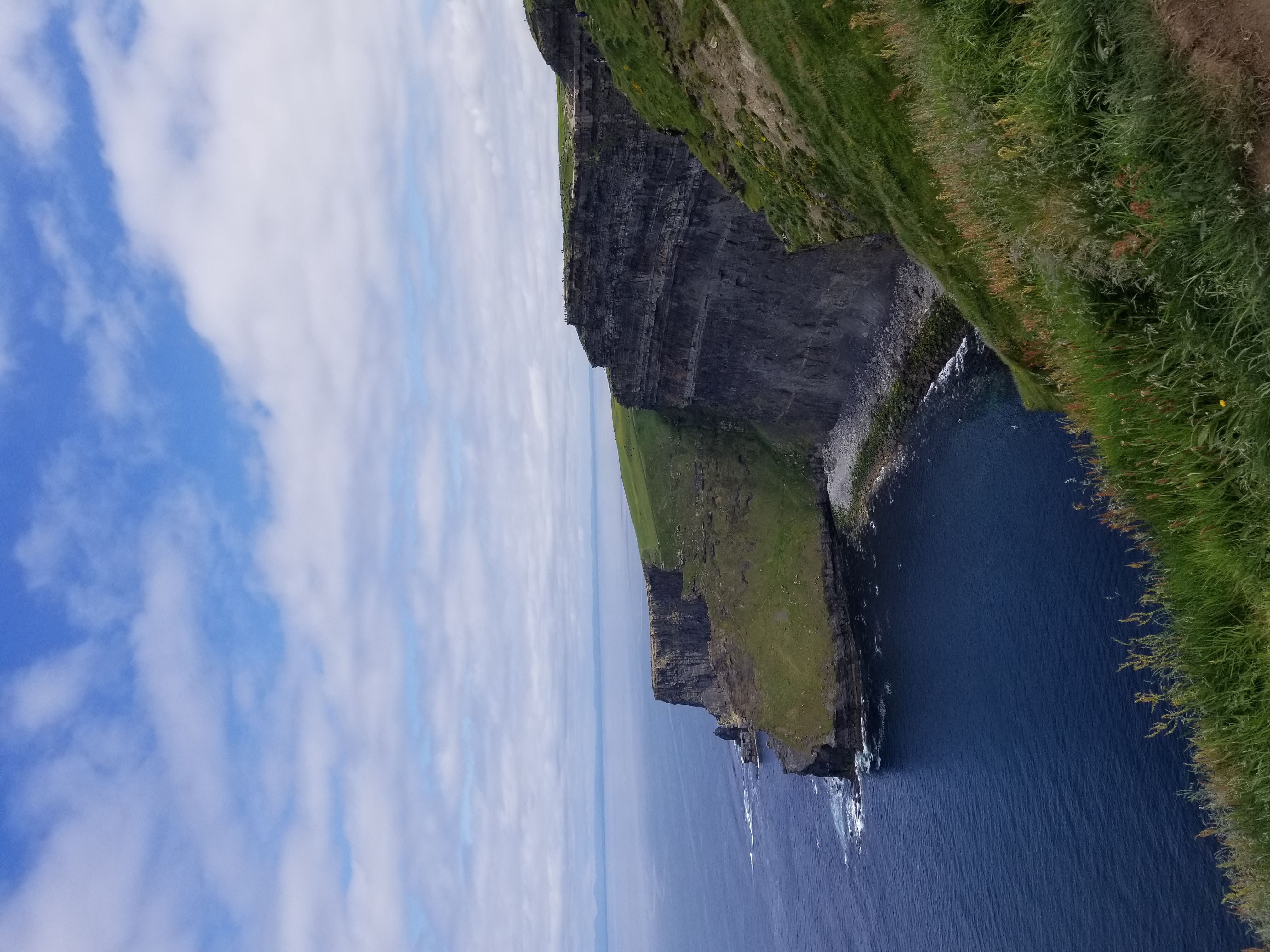 Cliff of Moher. A jagged cliff leading down into the ocean surrounded by sprawling green fields and a slightly cloudy, but otherwise blue, sky.