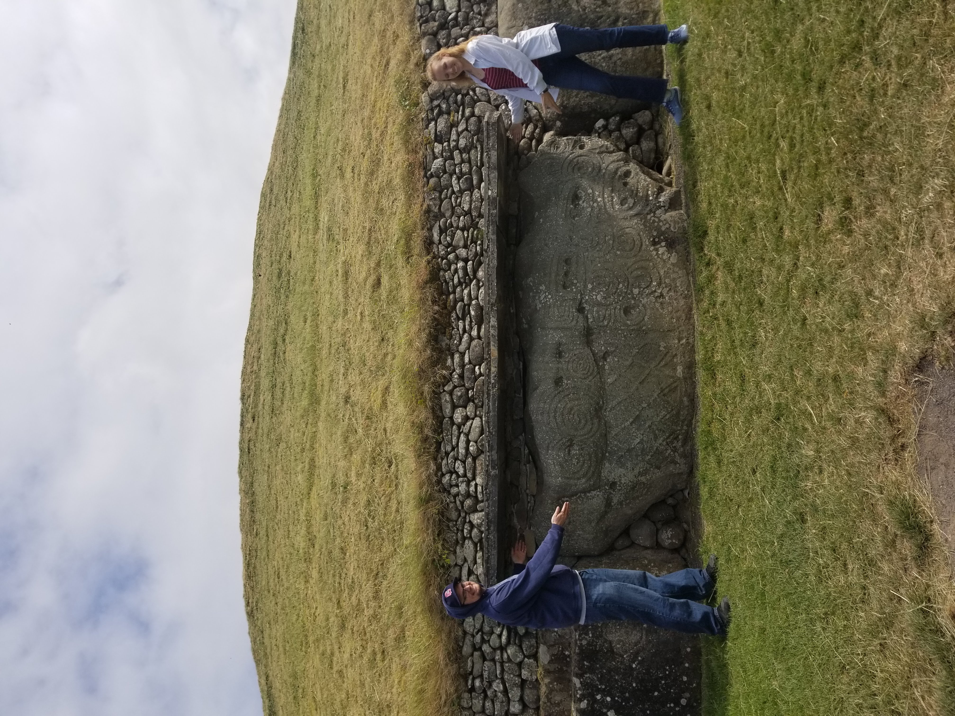 A kerbstone on the side of Newgrange, with smaller rocks positioned around it and the large grassy mound above. Author Jessica Dall and her partner stand on either side, showcasing it.