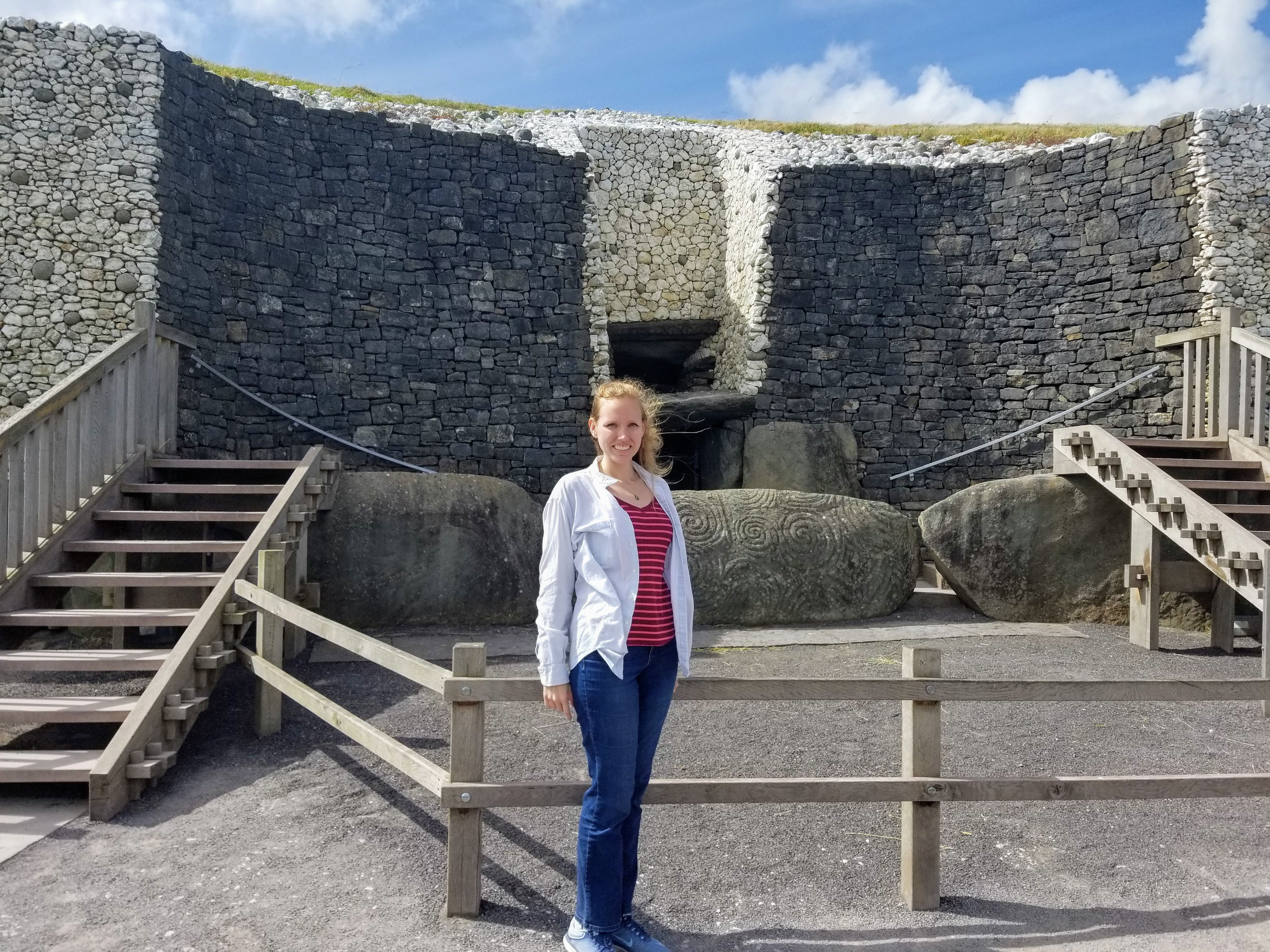 Newgrange. Large stone walls wrap around behind Author Jessica Dall. Immediately behind her are two sets of wooden steps leading to large stones, the one in the middle has been carved with megalithic art which curves in circles. The stone is called a kerbstone.