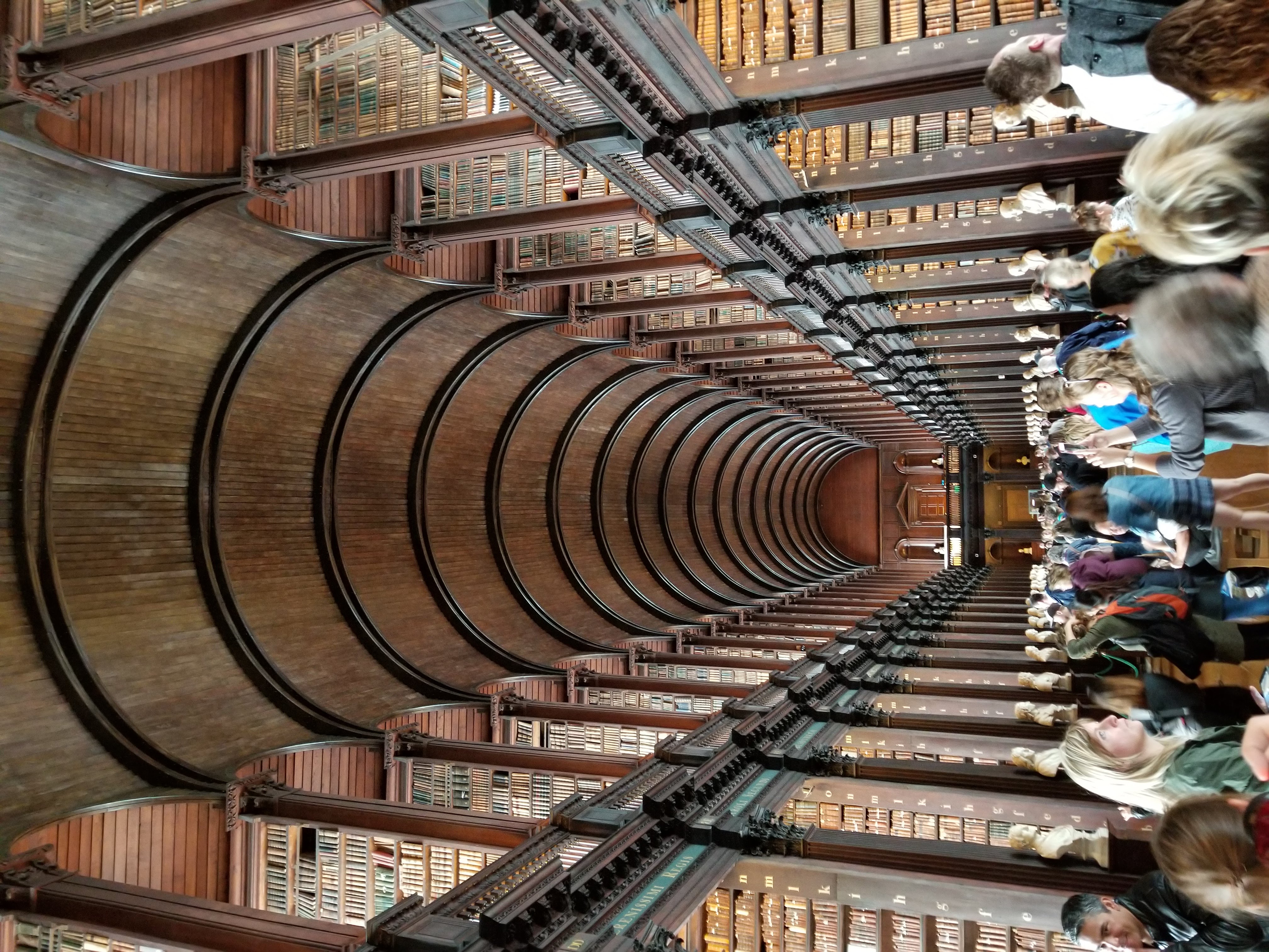 Trinity Library. A large arched ceiling, rows and rows of books on two separate floors, statues diving each aisle on the main floor. People are crowded along the walkway taking pictures.