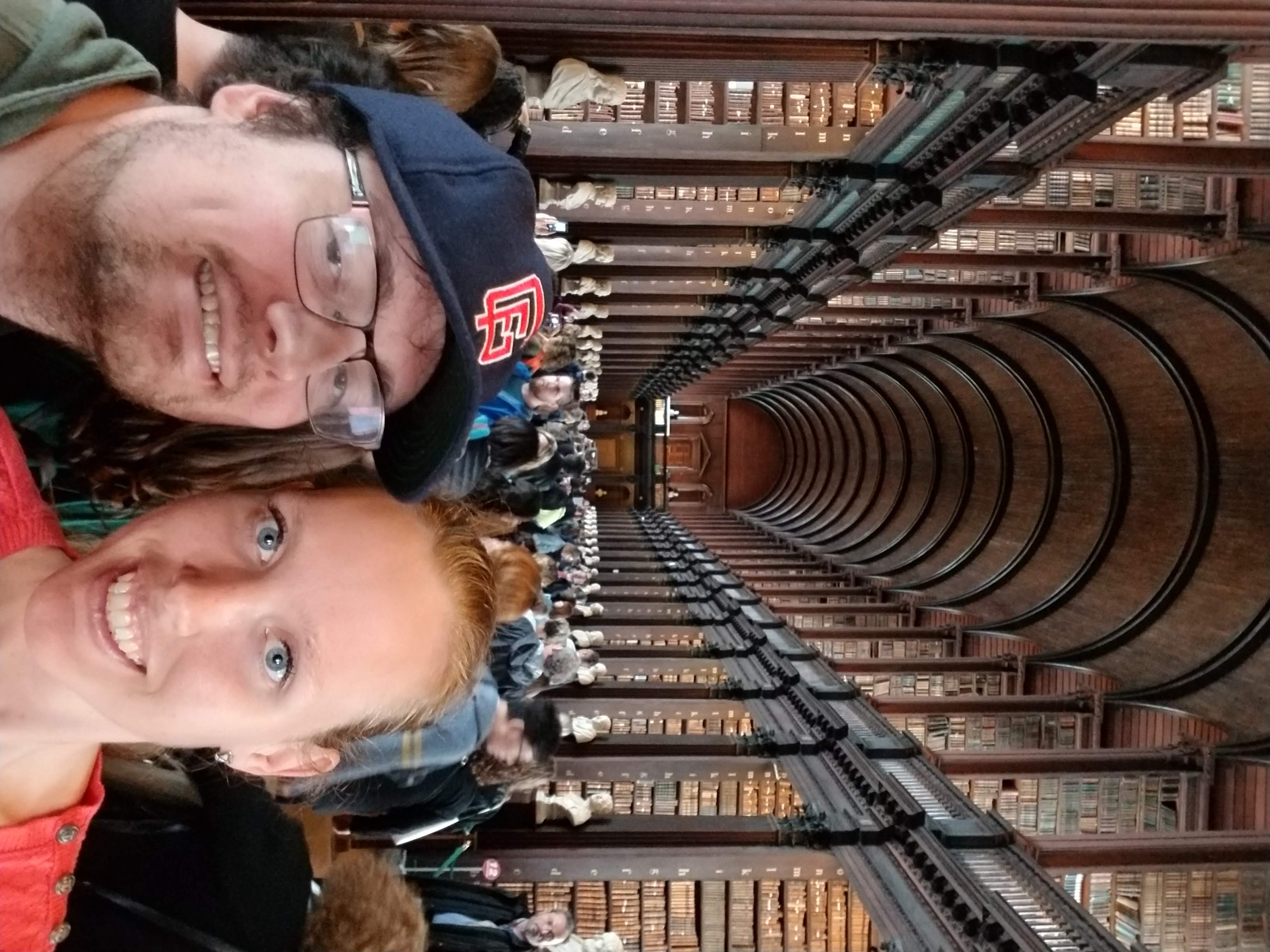Author Jessica Dall stands with her partner in Trinity Library. Behind them are rows and rows of books on two separate floors and a large arched ceiling above, with statues lining the lower walkway. Many people are walking behind them.