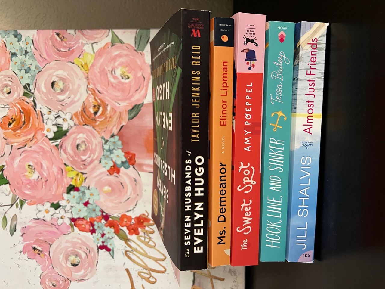 On a dark brown nightstand, five paperback books are stacked one on top of the other. Behind them is artwork of painted pink flowers in a pink vase.