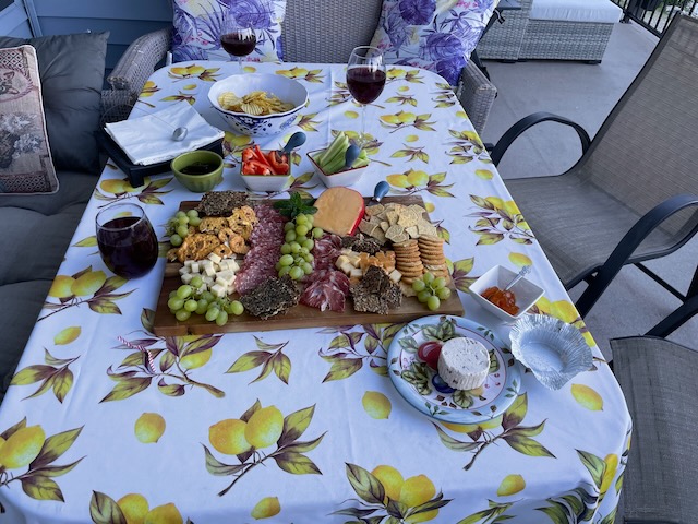 A charcuterie board, cheese, crudite, and glasses of wine on top of a lemon-printed table cloth