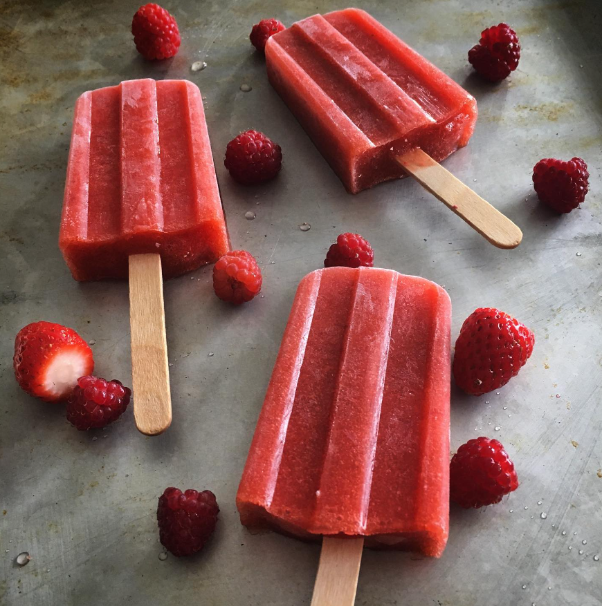 Three red-ish popsicles on wooden sticks laid flat on a countertop surrounded by raspberries and strawberries