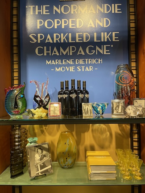 Sets of beautiful glassware spread across two shelves. Immediately behind, it says "The Normandie popped and sparkled like Champagne, Marlene Dietrich, Movie Star"